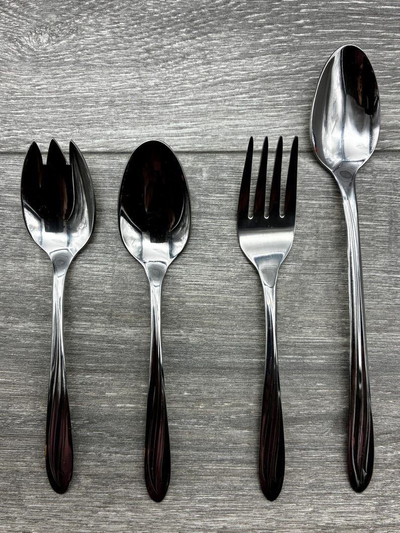 May include: Four-piece stainless steel flatware set. Includes a salad fork, a spoon, a dinner fork, and a serving spoon. The handles have a dark, reddish-brown color, and the utensils are arranged on a gray wooden surface.