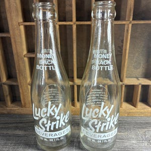 May include: Two clear glass Lucky Strike Beverages bottles. Each bottle has white text that reads "Lucky Strike Beverages" and "Money Back Bottle". Each bottle is 7 fl. oz.