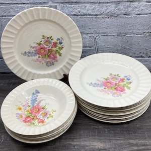 May include: A collection of off-white ceramic plates and bowls. Each piece features a floral design with pink, purple, and blue flowers. The plates have a ruffled edge, and the bowls are stacked.
