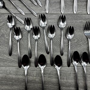 May include: An assortment of stainless steel silverware, including forks, spoons, and serving utensils, arranged on a gray wooden surface. The silverware has a shiny, reflective finish, and the arrangement suggests a formal dining setting.