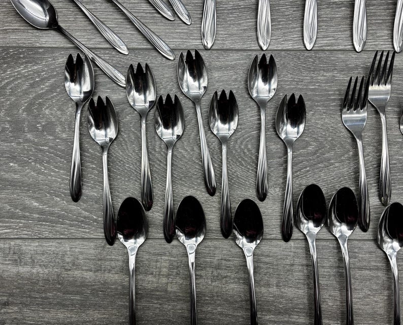 May include: An assortment of stainless steel silverware, including forks, spoons, and serving utensils, arranged on a gray wooden surface. The silverware has a shiny, reflective finish, and the arrangement suggests a formal dining setting.