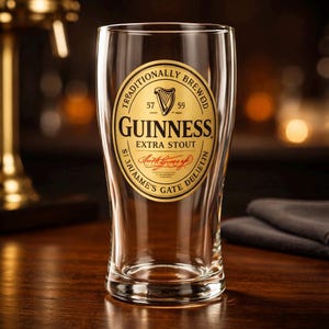 K&ouml;nnte beinhalten: Ein klares Pintglas mit dem Guinness-Logo. Das goldene Etikett hat schwarzen Text und ein Harfensymbol. Das Glas steht auf einer Holzoberfl&auml;che mit verschwommenem Hintergrund.