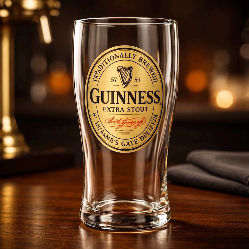 K&ouml;nnte beinhalten: Ein klares Pintglas mit dem Guinness-Logo. Das goldene Etikett hat schwarzen Text und ein Harfensymbol. Das Glas steht auf einer Holzoberfl&auml;che mit verschwommenem Hintergrund.