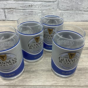 May include: Four clear glass tumblers with the Guinness Black Lager logo. The glasses have a gray and blue design with the Guinness harp logo and text. The glasses are on a wooden surface.