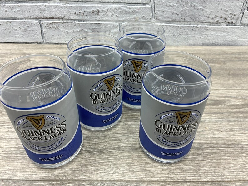 May include: Four clear glass tumblers with the Guinness Black Lager logo. The glasses have a gray and blue design with the Guinness harp logo and text. The glasses are on a wooden surface.
