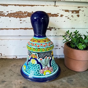 May include: A hand-painted ceramic bell with a dark blue handle and a flared base. The bell features colorful floral and geometric patterns in shades of blue, green, yellow, and red. The bell is approximately 6 inches tall.