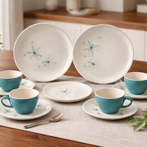 May include: A complete mid-century modern dinnerware set. The set includes large plates, saucers, and teal-colored cups. Each piece features a white background with a starburst design in teal and gold. A gold spoon rests on the table.