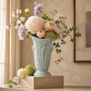 May include: A light blue ceramic vase with a textured design, filled with a variety of fresh flowers in shades of pink, white, and purple. The vase is displayed on a stone block, creating a visually appealing floral arrangement.