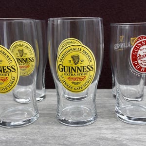 K&ouml;nnte beinhalten: Mehrere klare Pintgl&auml;ser mit dem Guinness-Logo. Die Gl&auml;ser haben ein gelb-schwarzes Etikett mit der Aufschrift "Guinness Extra Stout" und "St. James Gate Dublin". Ein Glas hat ein rot-wei&szlig;es Etikett mit der Aufschrift "Guinness Porter".