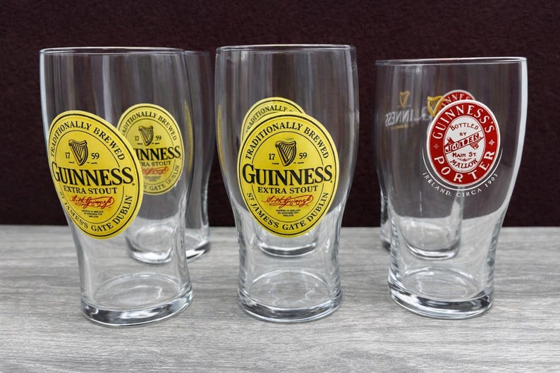 K&ouml;nnte beinhalten: Mehrere klare Pintgl&auml;ser mit dem Guinness-Logo. Die Gl&auml;ser haben ein gelb-schwarzes Etikett mit der Aufschrift "Guinness Extra Stout" und "St. James Gate Dublin". Ein Glas hat ein rot-wei&szlig;es Etikett mit der Aufschrift "Guinness Porter".