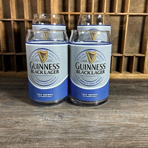 May include: Four clear Guinness Black Lager pint glasses. Each glass has a silver and blue label with the Guinness logo and text. The glasses are on a wooden surface with a wooden background.