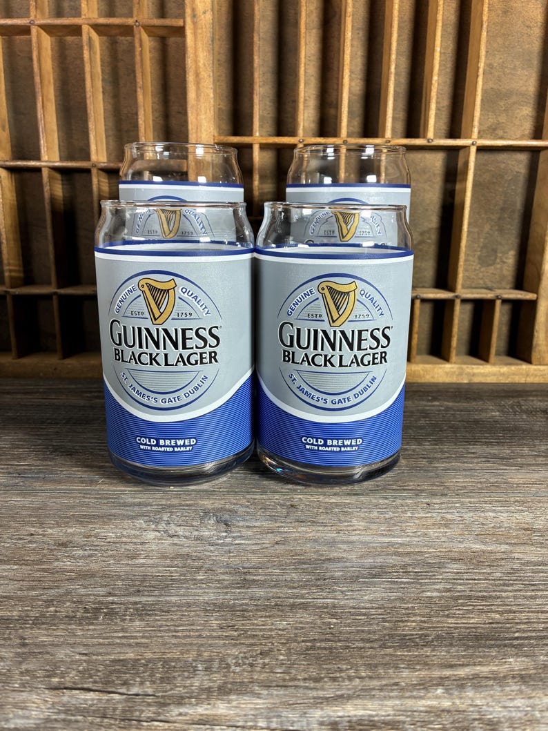 May include: Four clear Guinness Black Lager pint glasses. Each glass has a silver and blue label with the Guinness logo and text. The glasses are on a wooden surface with a wooden background.