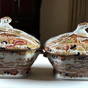 May include: Two antique covered serving dishes. Each dish features a detailed landscape scene with buildings, trees, and flowers in shades of brown, orange, and white. The lids have a decorative finial. The dishes are on a white surface.