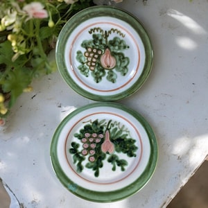 May include: Two white ceramic plates with green and pink floral designs. The plates feature a grapevine with grapes and a pear.