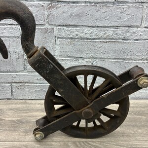May include: An antique, weathered black pulley system with a large curved hook. The pulley wheel has multiple spokes and is mounted within a metal frame. The metal shows signs of age and wear, suggesting it is an older piece.