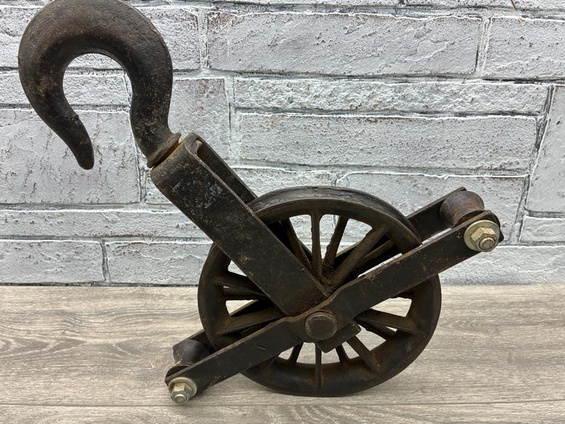 May include: An antique, weathered black pulley system with a large curved hook. The pulley wheel has multiple spokes and is mounted within a metal frame. The metal shows signs of age and wear, suggesting it is an older piece.