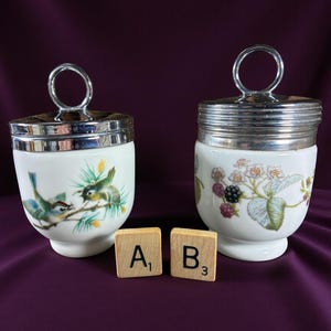 May include: Two white ceramic egg cups with silver-toned lids and handles. One cup features a bird and branch design, the other has a floral and berry pattern. Wooden letter tiles are placed in front of the cups.