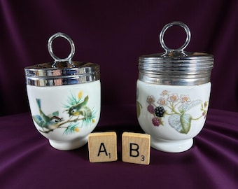 May include: Two white ceramic egg cups with silver-toned lids and handles. One cup features a bird and branch design, the other has a floral and berry pattern. Wooden letter tiles are placed in front of the cups.