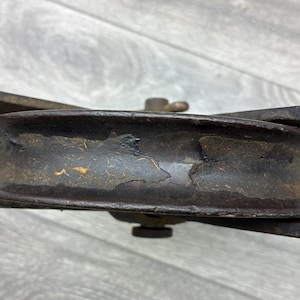 May include: Close-up of a vintage, weathered pulley wheel. The metal wheel is dark brown with areas of rust and wear, showing signs of age. The pulley has a central axle and is supported by two metal arms. The overall aesthetic suggests an antique or industrial item.