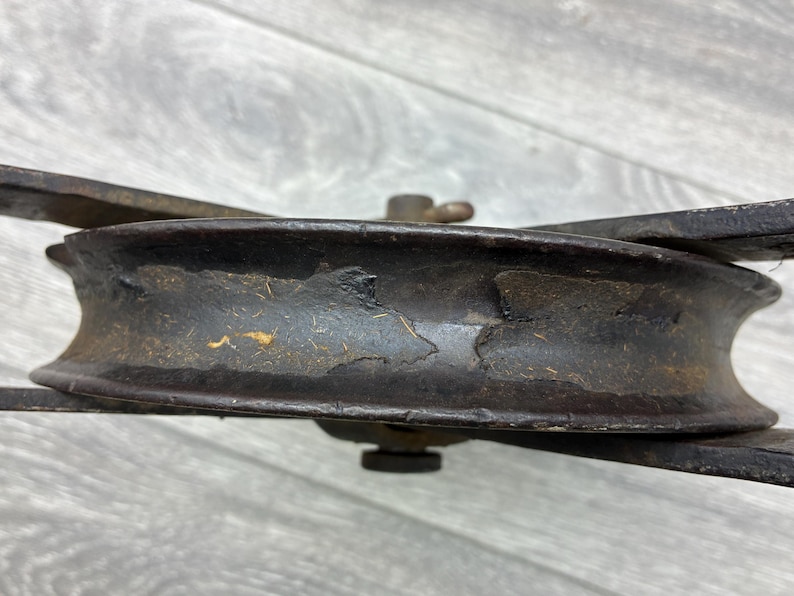 May include: Close-up of a vintage, weathered pulley wheel. The metal wheel is dark brown with areas of rust and wear, showing signs of age. The pulley has a central axle and is supported by two metal arms. The overall aesthetic suggests an antique or industrial item.