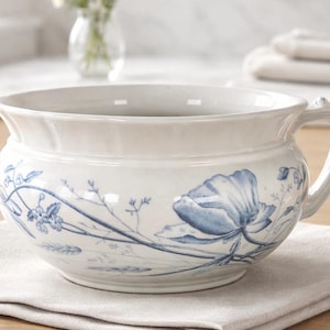 May include: White ceramic bowl with a handle, adorned with blue floral patterns. The bowl sits on a neutral-colored linen napkin. The design features a large flower and delicate botanical elements.