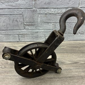 May include: An antique, cast iron pulley with a large hook and a spoked wheel. The pulley is dark gray with signs of wear and rust, suggesting age and use. The design is industrial and functional.