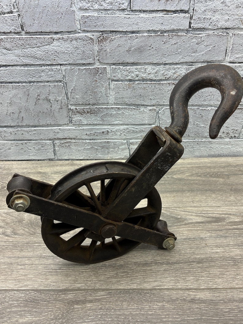 May include: An antique, cast iron pulley with a large hook and a spoked wheel. The pulley is dark gray with signs of wear and rust, suggesting age and use. The design is industrial and functional.