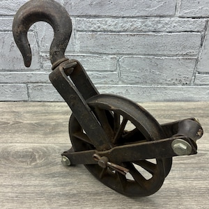 May include: An antique, rusty metal pulley with a large hook. The pulley has a wheel with spokes and is attached to a frame. The metal has a weathered appearance, suggesting age and use. The background is a light gray brick wall.