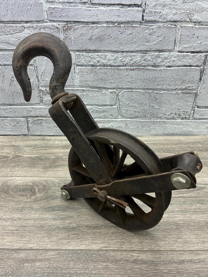 May include: An antique, rusty metal pulley with a large hook. The pulley has a wheel with spokes and is attached to a frame. The metal has a weathered appearance, suggesting age and use. The background is a light gray brick wall.