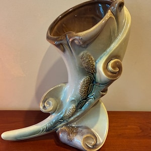 May include: A ceramic vase in a horn shape, with a light green and brown glaze. The vase features a curved design with embossed pinecone details and a curled accent. The vase is approximately 12 inches tall.