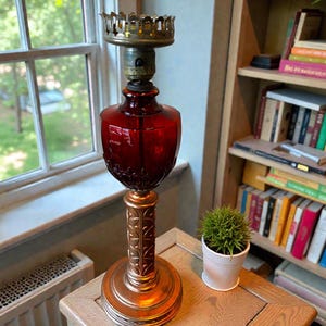 May include: A vintage table lamp with a ruby red glass shade and a decorative brass base. The lamp features a detailed, ornate design and stands approximately 18 inches tall. The lamp is placed on a wooden table near a window.