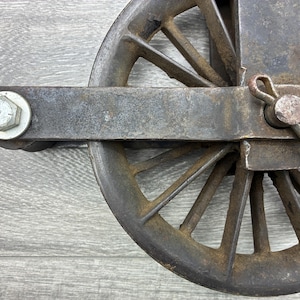 May include: Close-up of a weathered, antique metal pulley wheel. The wheel has multiple spokes and is attached to a rectangular metal frame with a bolt and pin. The metal has a rusty, aged appearance.
