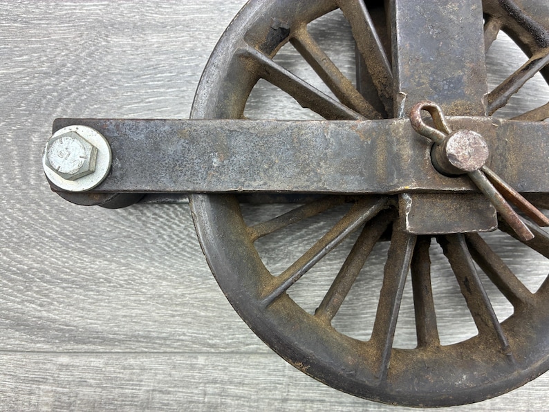 May include: Close-up of a weathered, antique metal pulley wheel. The wheel has multiple spokes and is attached to a rectangular metal frame with a bolt and pin. The metal has a rusty, aged appearance.
