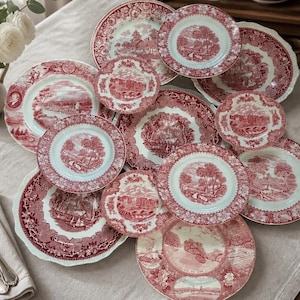 May include: A collection of ceramic plates featuring a red and white transferware pattern with detailed landscape scenes and floral borders. The set includes various sizes of round plates, some with scalloped edges, arranged in a layered display.