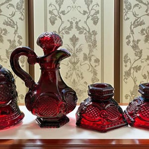 May include: A collection of four ruby red glass decorative items. The set includes a bell-shaped bottle, a pitcher with a handle, and two square-based containers. All pieces feature ornate, embossed patterns.