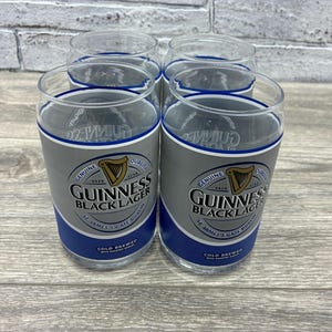 May include: Four clear pint glasses with a blue and silver band featuring the Guinness Black Lager logo. The glasses have a classic pint shape and are designed for enjoying cold beverages. The glasses are displayed on a light-colored wooden surface.