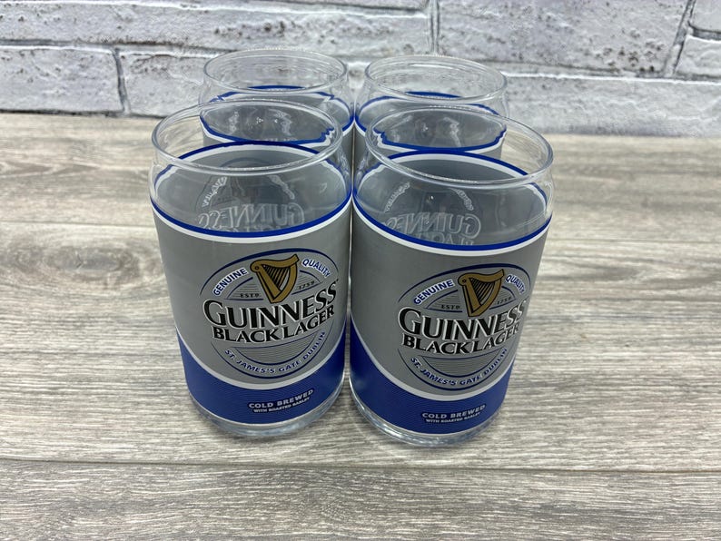 May include: Four clear pint glasses with a blue and silver band featuring the Guinness Black Lager logo. The glasses have a classic pint shape and are designed for enjoying cold beverages. The glasses are displayed on a light-colored wooden surface.