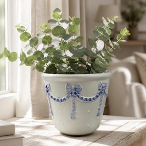 May include: A white ceramic planter featuring a raised blue relief pattern of draped swags and bows around the upper rim, with small white raised dots across the body, containing a green eucalyptus plant with rounded leaves and thin stems.