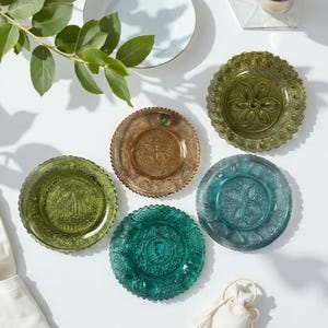 May include: A collection of decorative glass plates in various colors, including green, teal, and amber. Each plate features a unique embossed design and a scalloped edge. The plates are arranged on a white surface.
