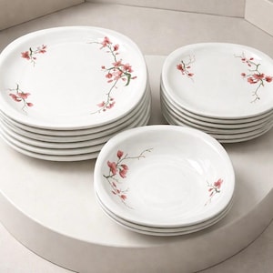 May include: A set of white ceramic dinnerware, including plates and bowls, arranged on a white surface. Each piece is decorated with a delicate floral design in shades of red and pink, adding a touch of elegance to the set.