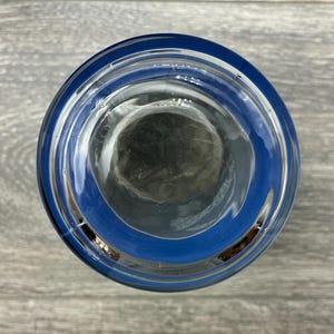 May include: A clear glass with a blue rim, viewed from above. The glass is empty, and the blue rim creates a distinct border around the opening. The glass sits on a gray wooden surface.