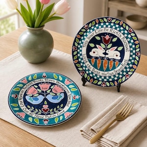May include: Two round ceramic decorative plates featuring colorful folk-art style illustrations. One plate displays two white rabbits and carrots with the text "May Your Life Be Richly Blessed With The Best The Sun Brings Forth". The other shows two blue birds with the text "Love Rejoices, Trusts, Hopes, Perseveres, Never Fails. Over All These Put On Love".