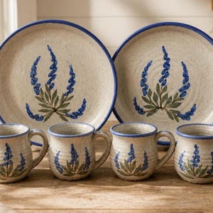 May include: A set of two round ceramic plates and four matching mugs, all featuring a speckled cream finish with a blue rim and a hand-painted blue lupine flower design with green leaves centered on each piece of dinnerware.