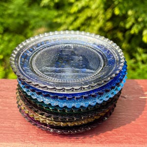 May include: A stack of colorful glass coasters in shades of blue, green, and purple. Each coaster has a detailed embossed design and a scalloped edge. The coasters are stacked on a red wooden surface.