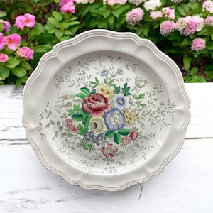 May include: A white ceramic plate with a scalloped edge features a colorful floral design. The plate is decorated with a bouquet of pink, red, yellow, blue, and purple flowers with green leaves.
