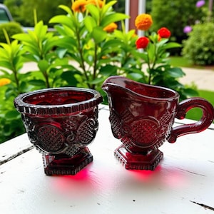 May include: A ruby red glass creamer and sugar bowl set. The creamer has a handle and spout, while the sugar bowl is open. Both pieces have a detailed, textured pattern and sit on a pedestal base. The set is displayed on a white surface with a green background.