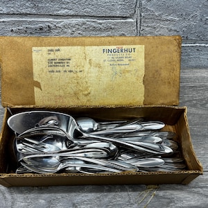 May include: A cardboard box filled with stainless steel spoons and serving utensils. The box has a faded label with text from Fingerhut Products Co. The spoons are stacked inside the open box, reflecting light.
