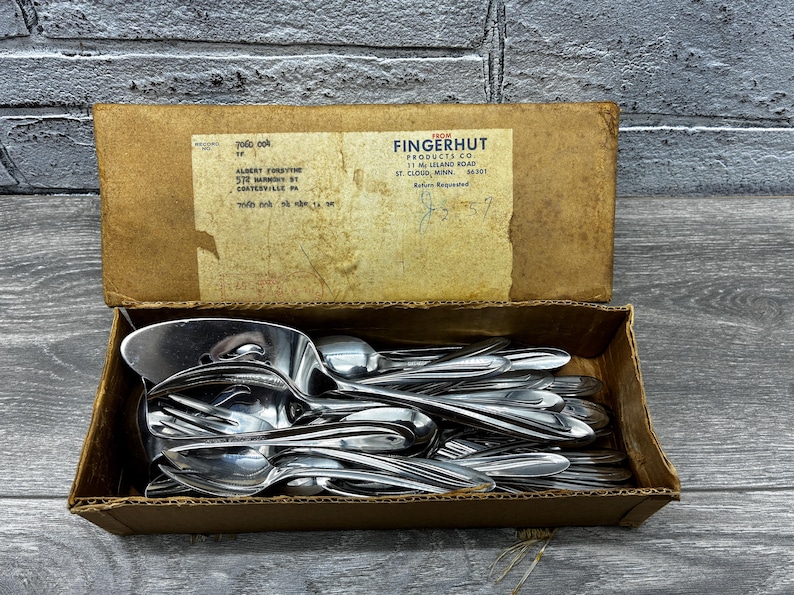 May include: A cardboard box filled with stainless steel spoons and serving utensils. The box has a faded label with text from Fingerhut Products Co. The spoons are stacked inside the open box, reflecting light.