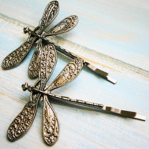 Pode incluir: Dois grampos de cabelo de libélula prateados com intrincados desenhos florais. Os grampos estão sobre um fundo de madeira azul.