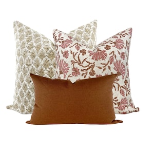May include: Three decorative throw pillows with a variety of patterns. The pillows are arranged in a stack, with the top pillow featuring a floral design in shades of pink and brown on a white background. The middle pillow has a geometric pattern in shades of gold and white. The bottom pillow is a solid rust color.